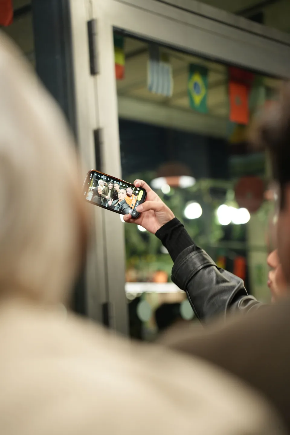 A blurred shoulder in the foreground; at the centre, a hand holds up a smartphone to photograph the room beneath hanging flags.