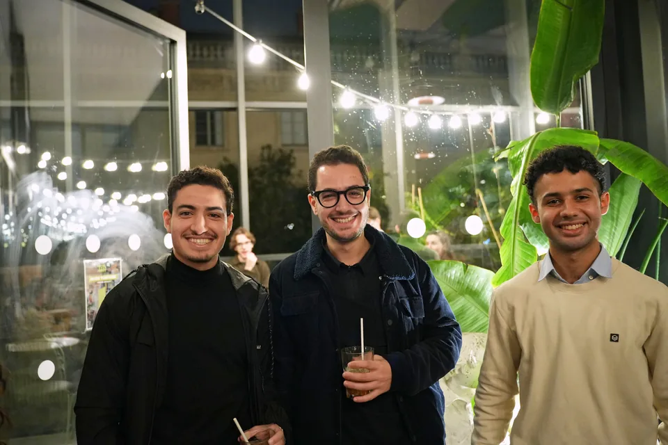 Three young men smile at the camera in front of a glass wall, strings of lights and a banana plant.