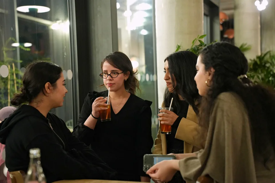 Four young women talk around a table, drinks in hand, in warm light.