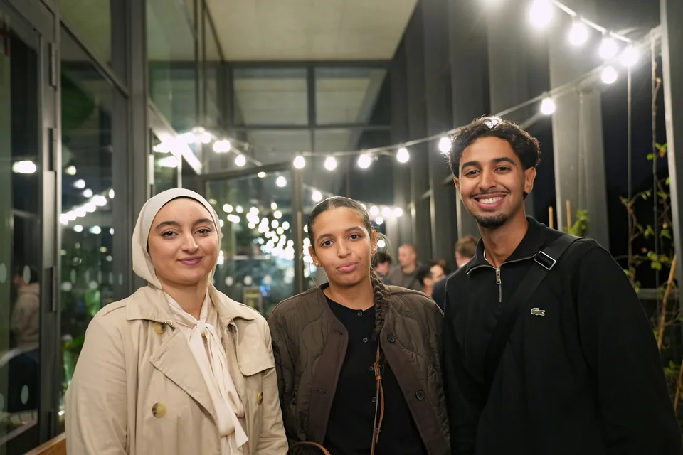 Three guests pose in a glass-walled gallery lined with strings of lights receding into the distance.