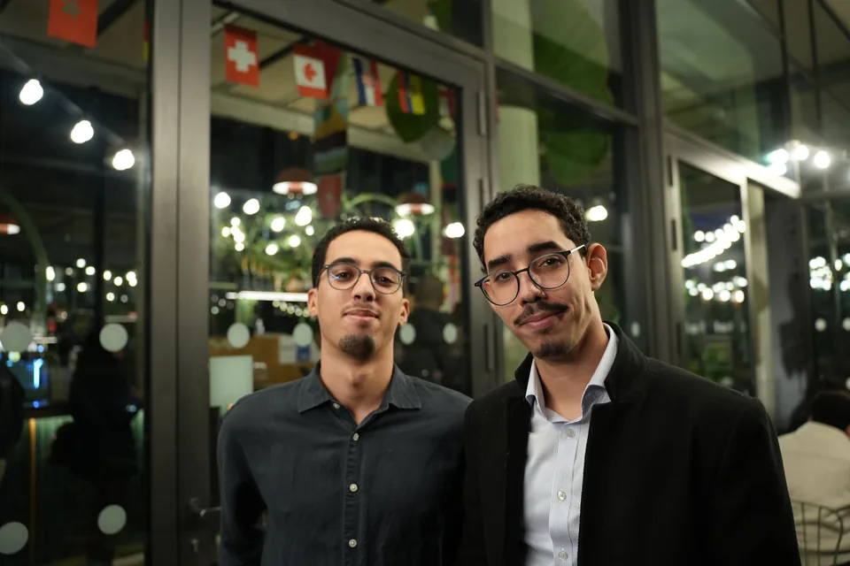 Two young men in glasses pose in front of a window decorated with flags of several countries.