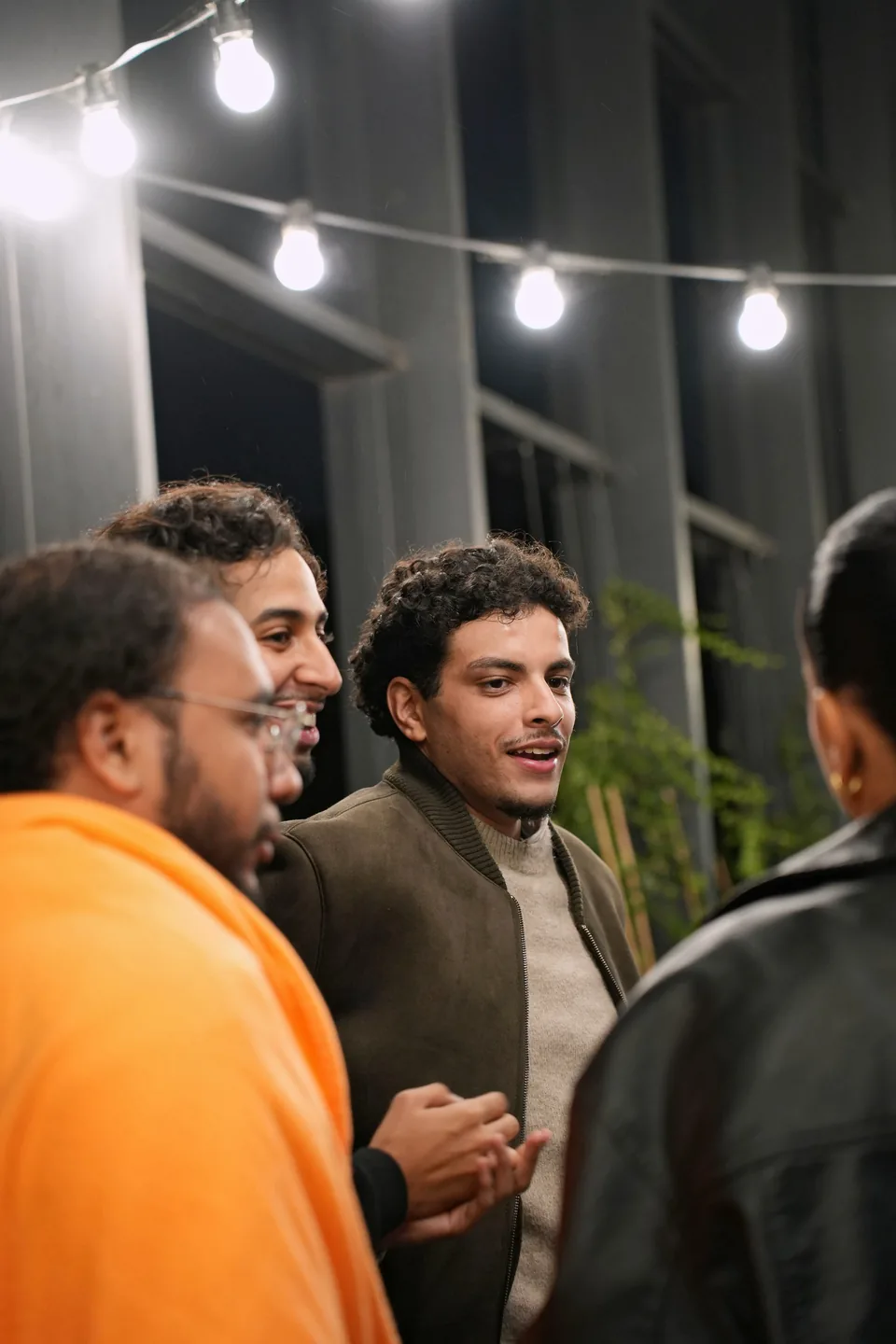 A group talks beneath strings of bulbs; a man in an orange hoodie in the foreground, another smiling at the centre.