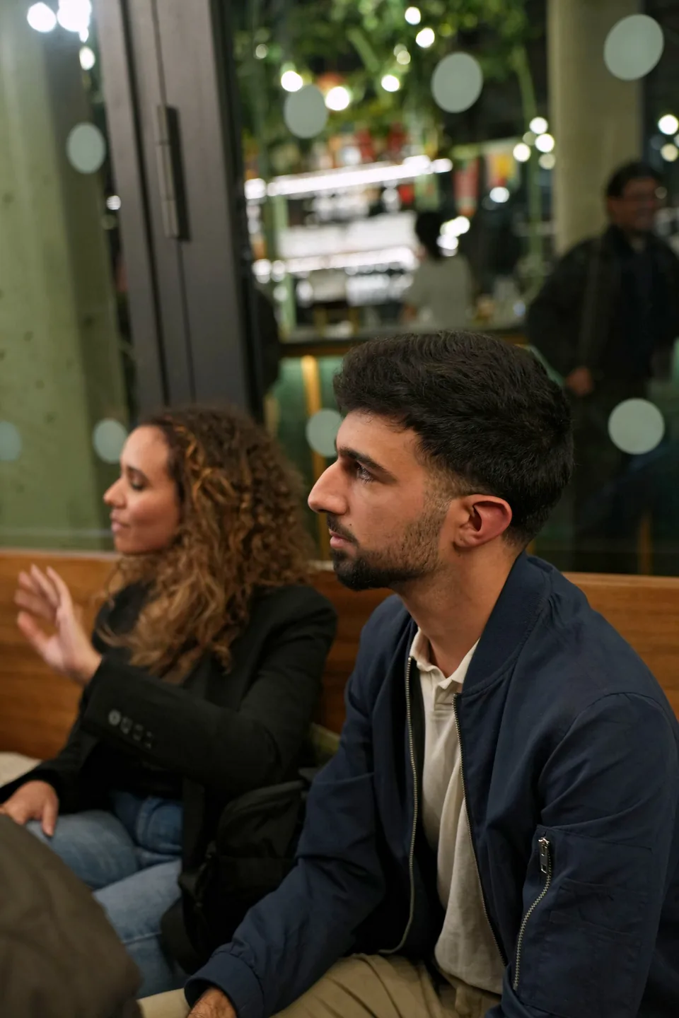 A woman with curly hair speaks with an expressive gesture while a young man in a blue jacket listens in profile.