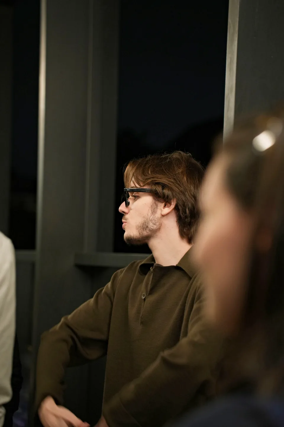 A young man with glasses in a khaki polo shirt listens in profile, a blurred face in the foreground.