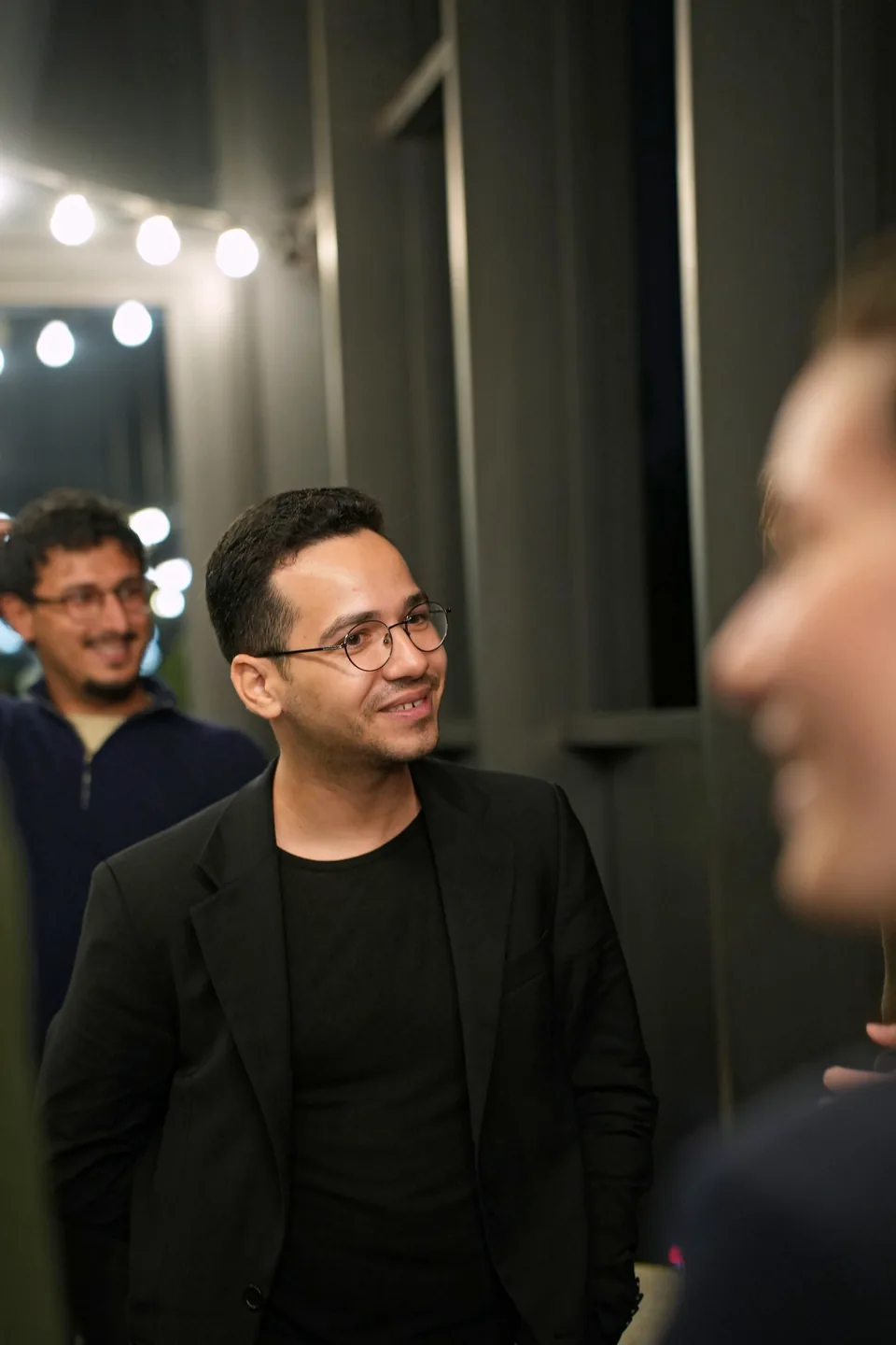 A man with round glasses and a black jacket smiles beneath strings of lights; another guest smiles behind him.