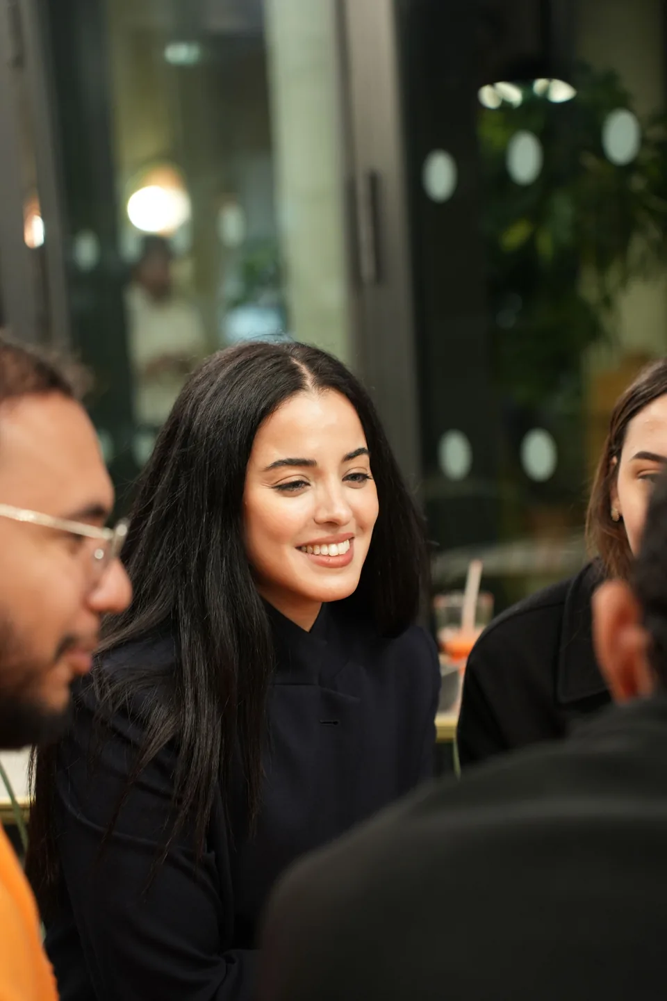 A young woman with long dark hair smiles among other guests, soft lights glowing behind her.