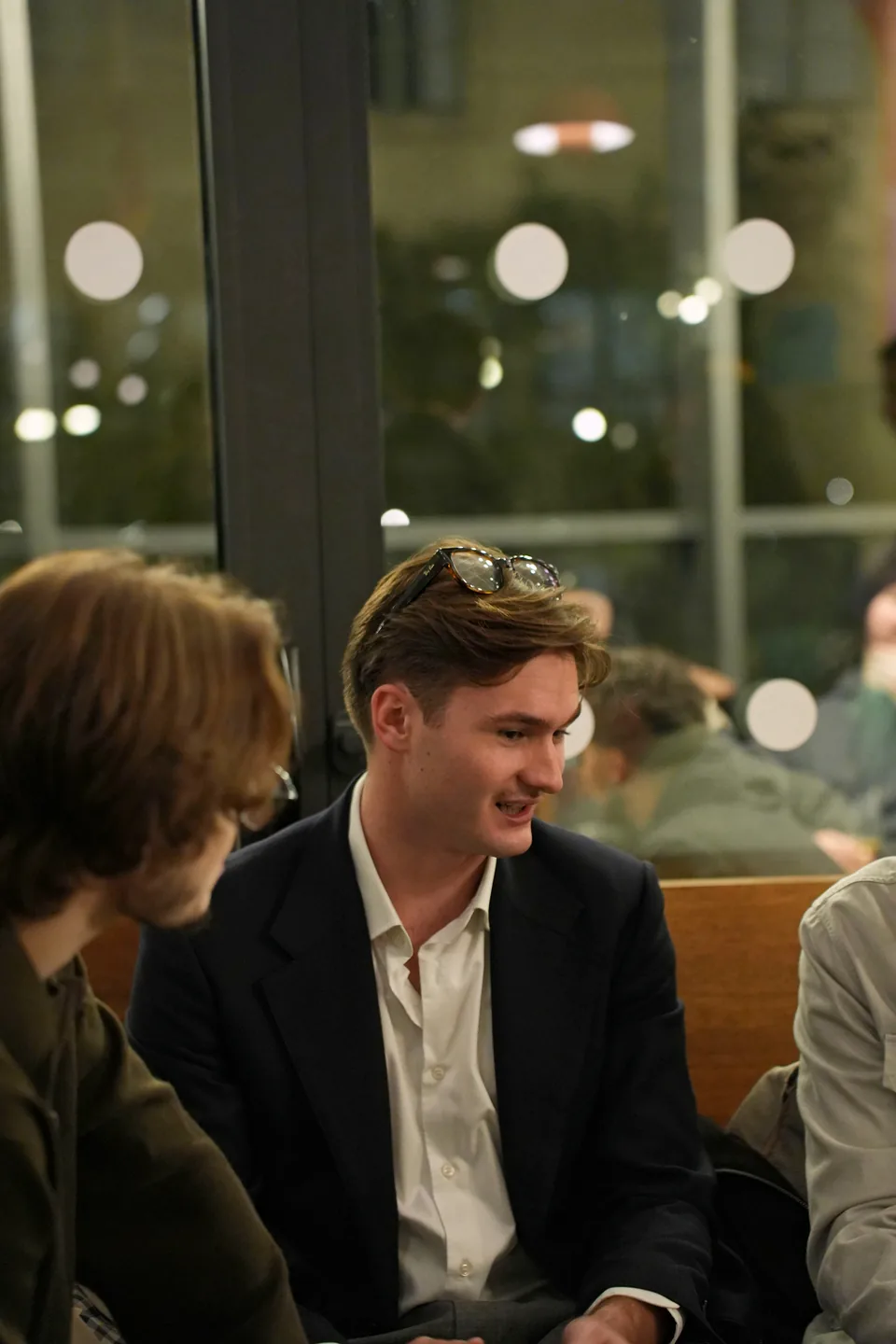 A young man in a blazer, sunglasses on his head, smiles as he listens to the person beside him on a banquette.
