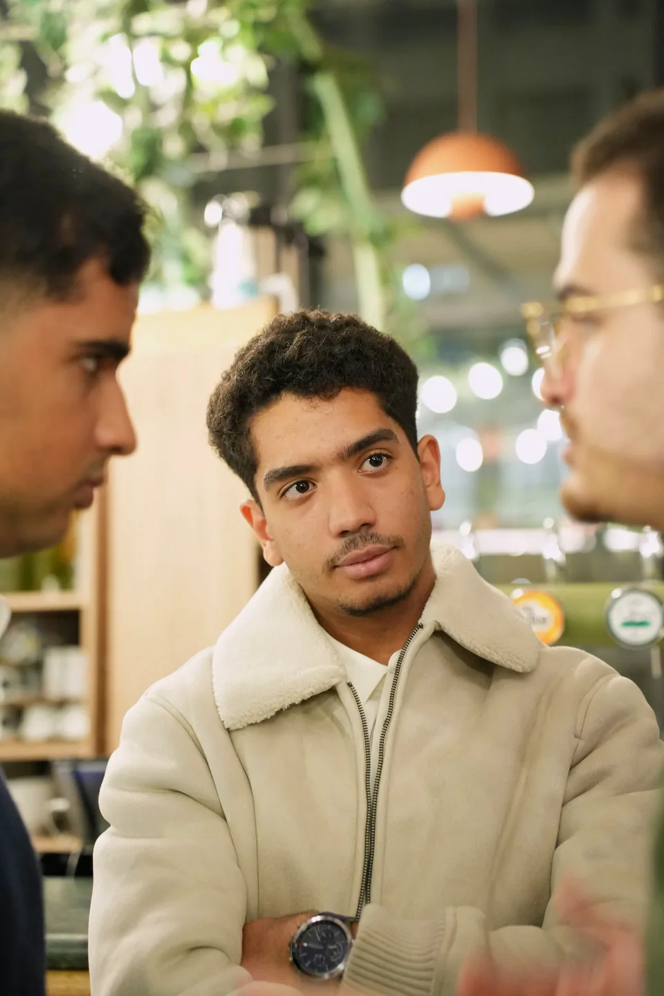 A young man in a shearling jacket listens with his arms crossed, framed by two blurred figures.