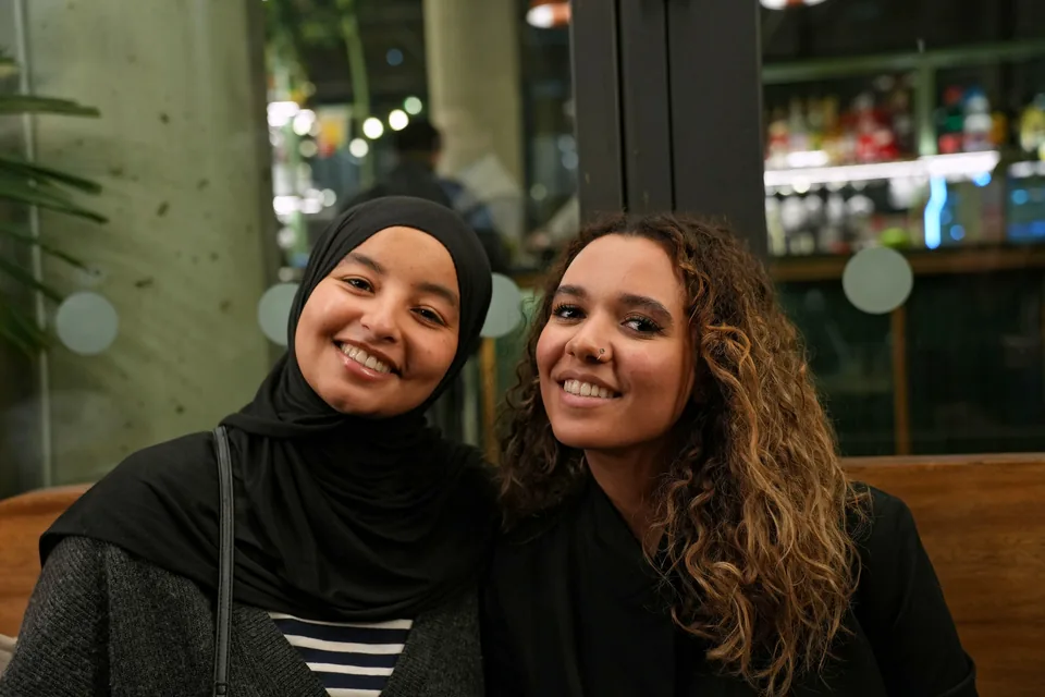Two young women, one wearing a black headscarf, smile side by side on a banquette.